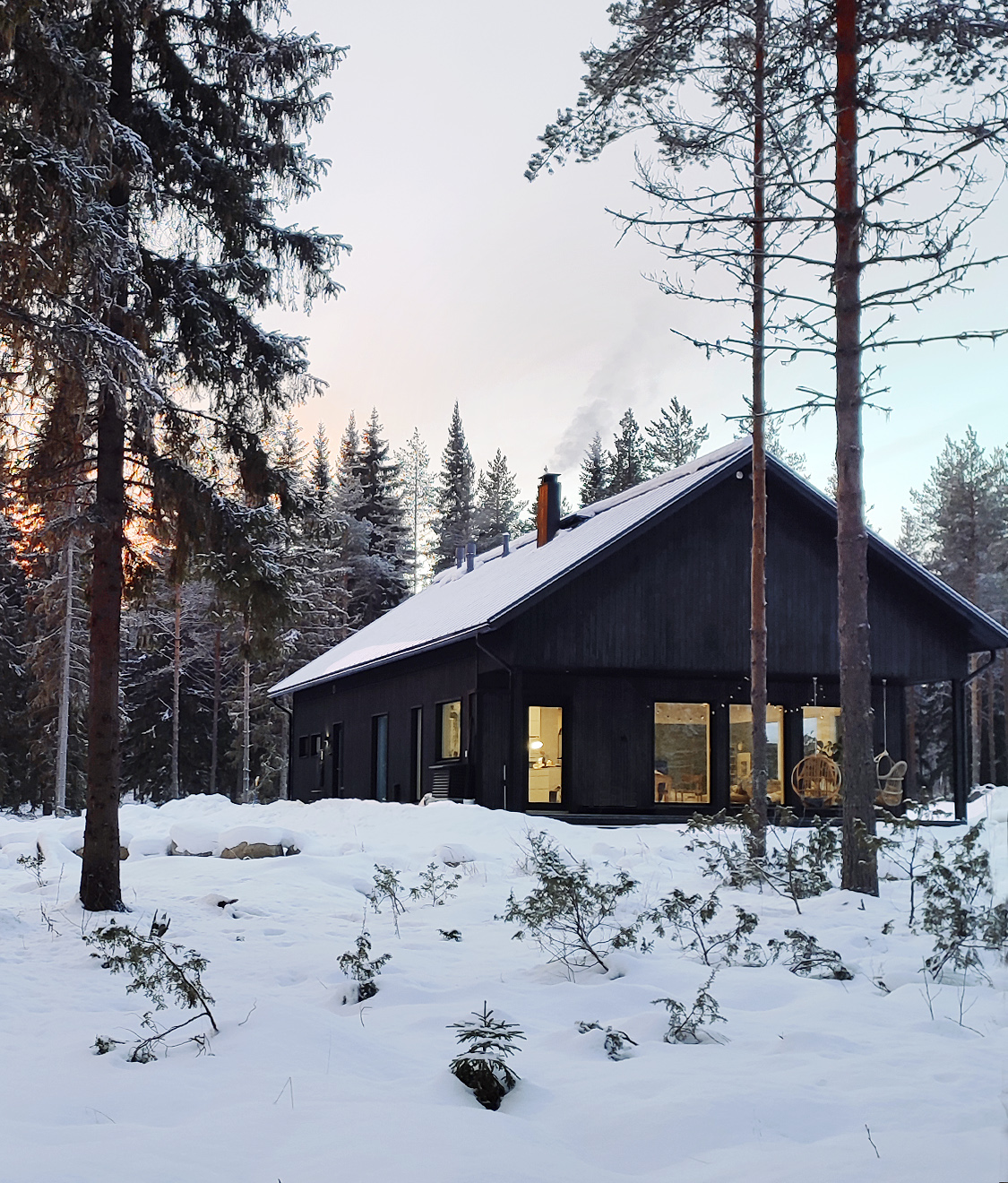 Musta yksikerroksinen talo korkealla harjalla talvisessa metsämaisemassa.